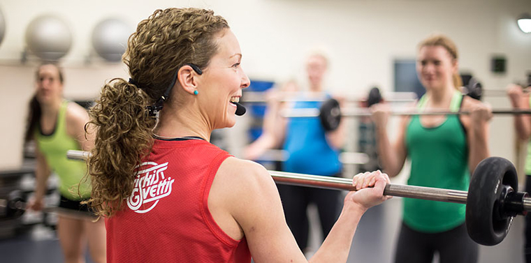Kvinna med lockigt hår och rött friskis-linne instruerar tränings-deltagare i skivstång.