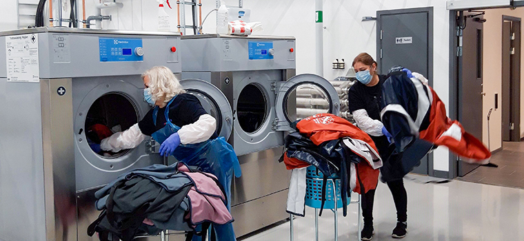 Två kvinnor med munskydd och förkläden matar in stora mängder tvätt i två stora tvättmaskiner.
