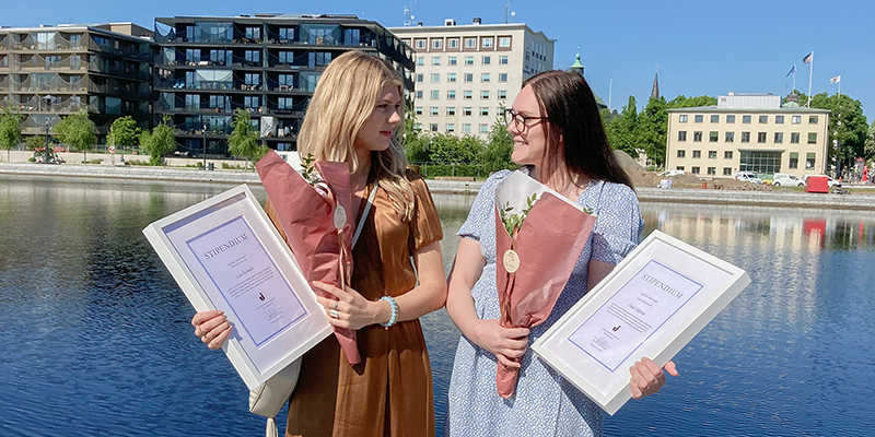 Stipendiaterna Frida och Lisa håller i sina stipendie-diplom och blombuketter. i bakgrundens syns vatten, en blå himmel och en stadsmiljö.