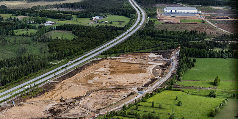 En stor väg (E4an) slingrar sig igenom grön mark och skog samt jordfärgad, delvis obebyggd industrimark.